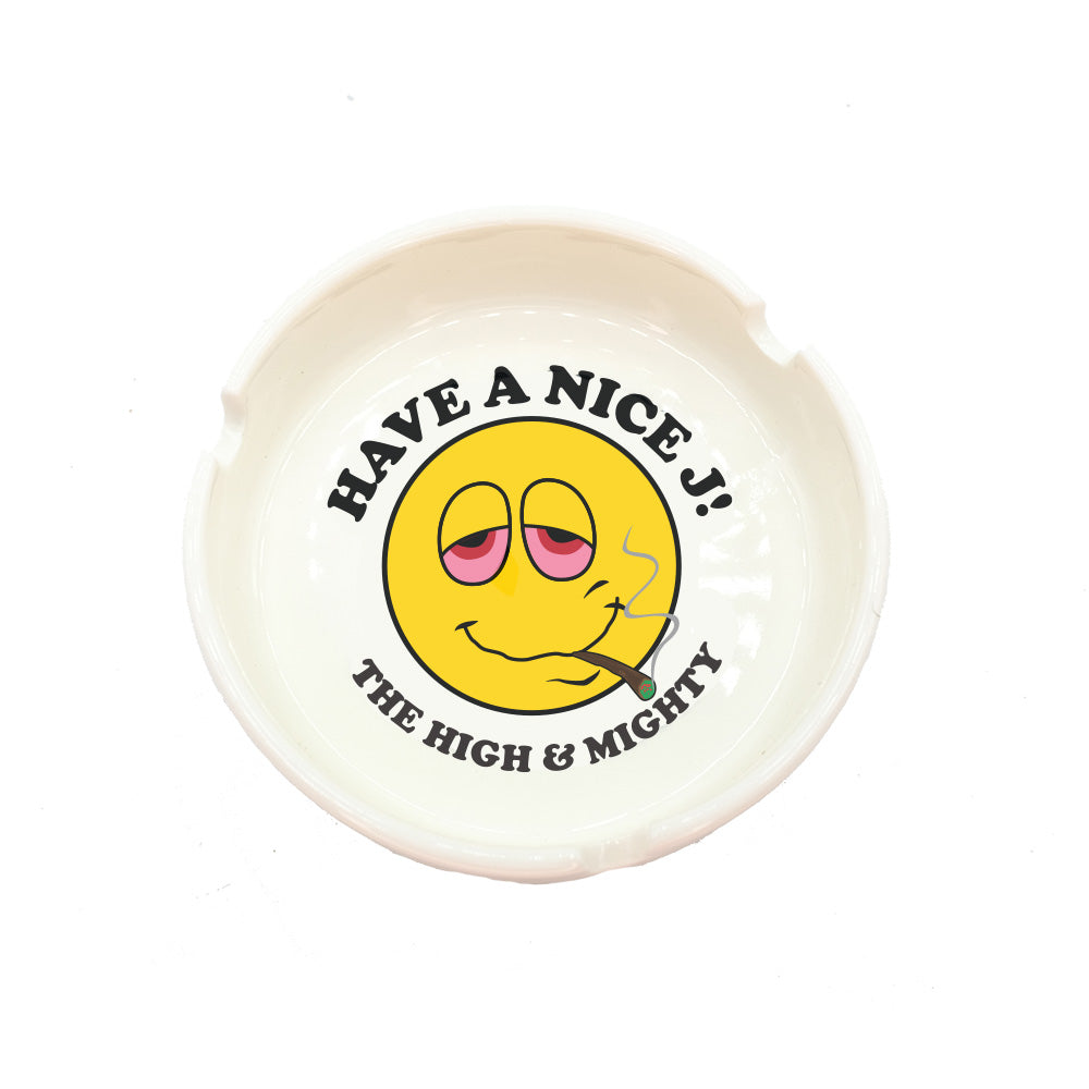 White ceramic ashtray with cartoon smile face with joint in mouth. Around picture it reads have a nice j!